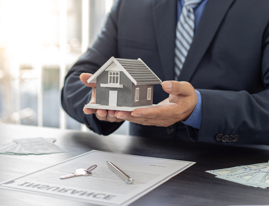 Man's hands holding a grey house over an insurance document with a key and pen.